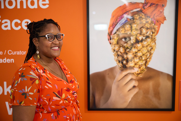 A smiling woman standing beside a photograph of a person with a scarf on their head and a part of a pineapple covering her face and just showing her eyes.