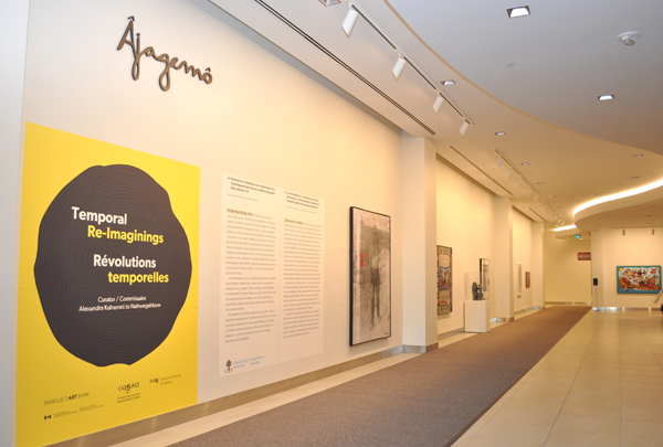 A gallery space with "Âjagemô" written in cursive above a large graphic and text panels and artworks installed on the walls further down in the space.