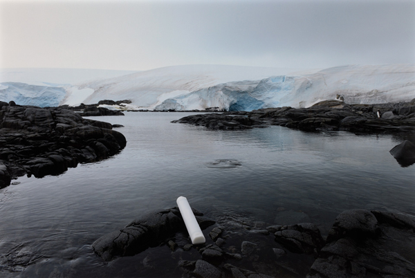 A white cylindrical tube lies on a rocky surface at the edge of water. Icebergs are in the backdrop.