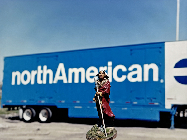 In the background is a blue tractor trailer with "northAmerican." written across it. In the foreground is plastic figurine of an Indigenous chief.