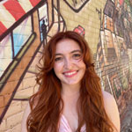 Photo of a young woman with long wavy auburn hair standing outside in front of a grafitti wall