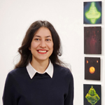 A headshot of a smiling woman with long black hair stands next to four small paintings installed vertically on a wall