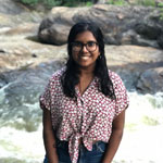 A young woman with brown skin and black hair and dark glasses is standing in front of a backdrop of sloping smooth rocks and forest