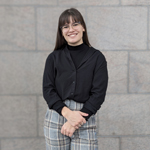 A woman with long brown hair, bangs and glasses wearing a black buttoned up sweater and grey plaid pants.