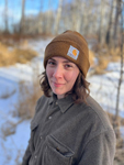 A headshot of a woman wearing a brown hat and grey jacket in a forest with snow on the ground.