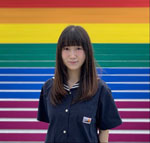 A headshot of an asian woman with shoulder-length dark brown hair standing in front a backdrop made up of bright horizontal colourful stripes.